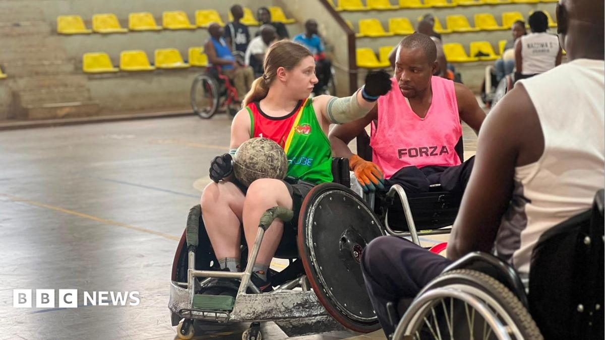 Sarah Burrows (left) in conversation with a man in a sports hall. They are both in wheelchairs. Sarah has a round ball in her lap. She has brown hair, tied back, and is wearing a red and green top. The man next to her has short black hair and is wearing a pink top.
