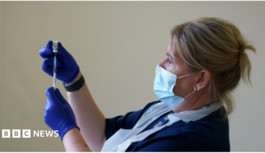 A blonde nurse with protective coverings on her face, hands and chest holds up a needle and vaccine vial.