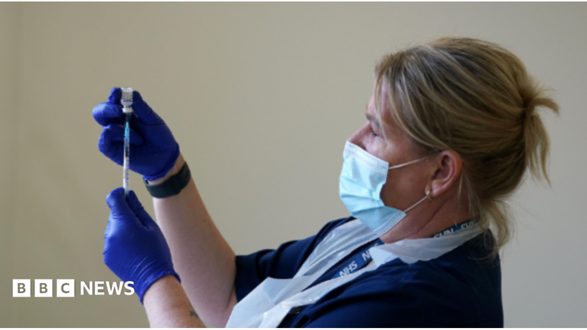 A blonde nurse with protective coverings on her face, hands and chest holds up a needle and vaccine vial.