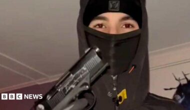 A teenager wearing a black balaclava and hoodie holding up a handgun, with his gloved hand on the trigger