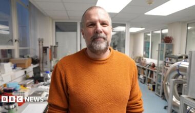 Richard wears a dark blue shirt which is unbuttoned at the collar. He is looking towards the camera with several screens showing images of the sun behind him