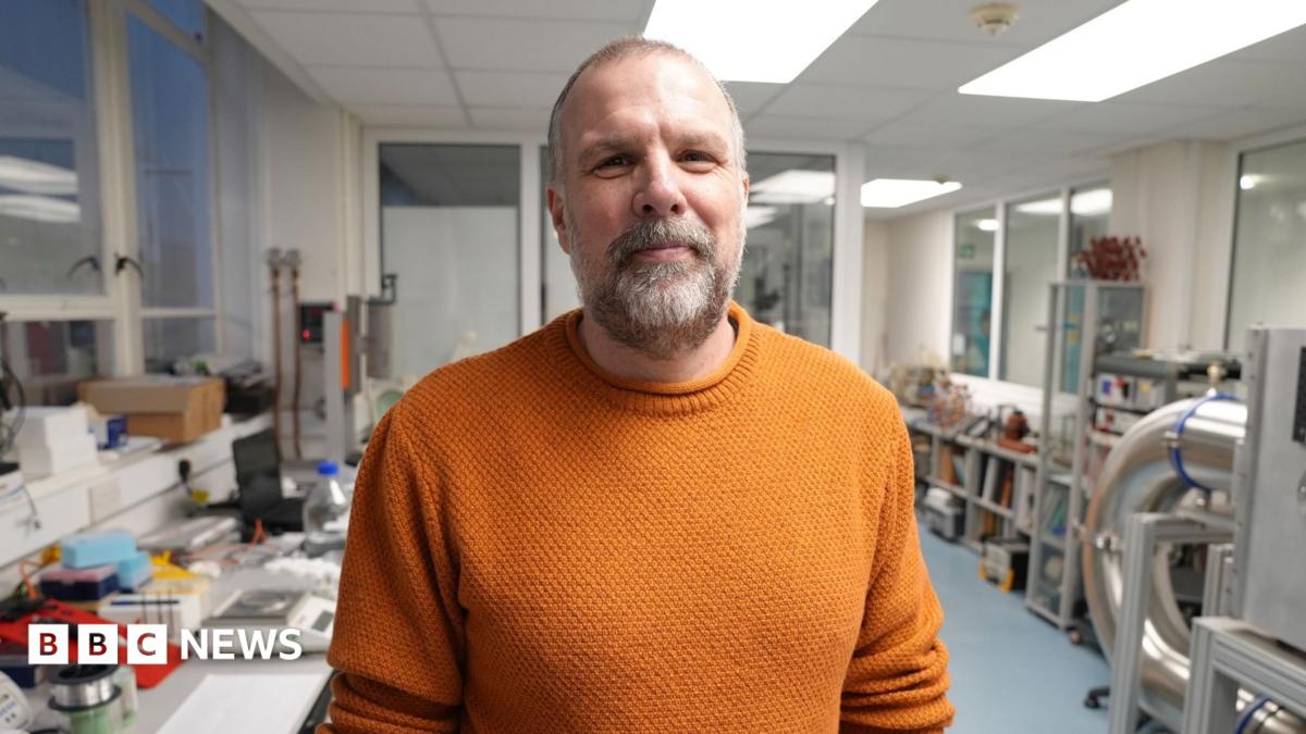 Richard wears a dark blue shirt which is unbuttoned at the collar. He is looking towards the camera with several screens showing images of the sun behind him