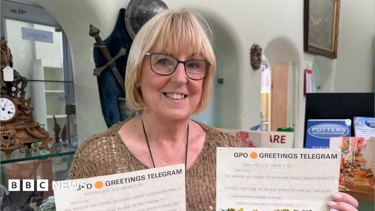 A woman smiles at the camera. She is holding two pieces of paper which have the headers "GPO Greetings Telegram". She is standing in front of multiple archways. There is a wooden clock on the left, and a sword is hung up on the wall.