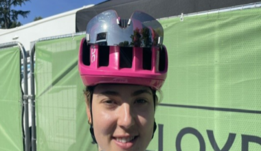Alice Towers wearing a pink cycling helmet with sports sunglasses attached. She is wearing a white cycling jersey and stood in front of a green fence.