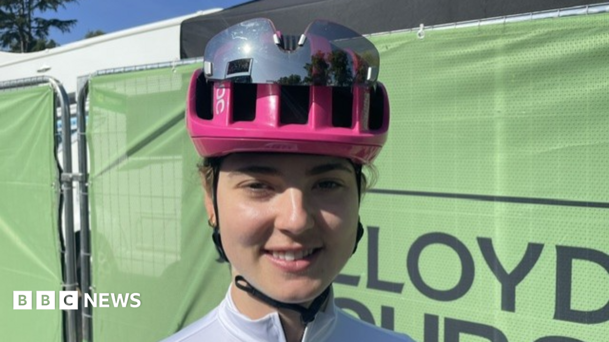 Alice Towers wearing a pink cycling helmet with sports sunglasses attached. She is wearing a white cycling jersey and stood in front of a green fence.