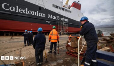Workers make final preparations ahead of the launch of the MV Glen Rosa in April 2024. Six workers can be seen in the foreground with their back to the camera. The white, black and red ferry, with Caledonian MacBrayne written on the side, is in the distance.