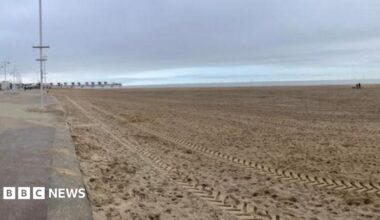 A view of the beach on a cloudy day.  A vehicle has left tracks in the sand in the foreground. Part of the promenade is in the left hand side of the picture.  The beach is deserted apart from a couple walking their dog in the distance.