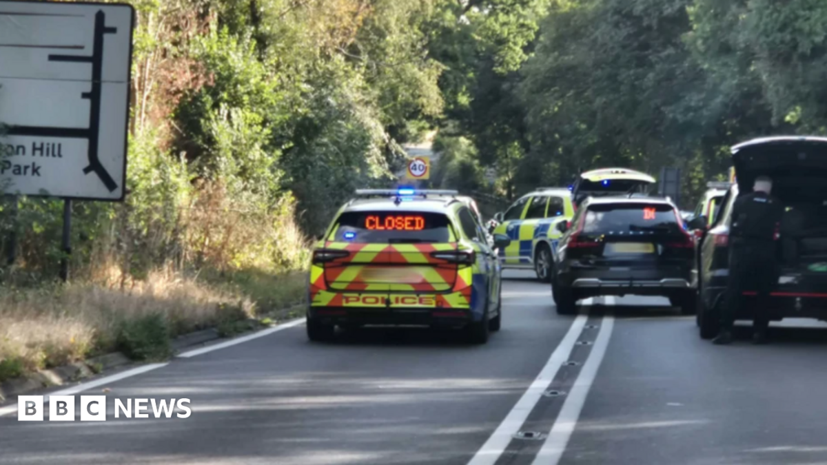Police cars can be seen in the road with other unmarked vehicles blocking the route. There is a road sign on the left with trees lining the road.