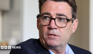 Andy Burnham is wearing a dark-coloured jacket, light blue shirt and glasses. He has short black hair.