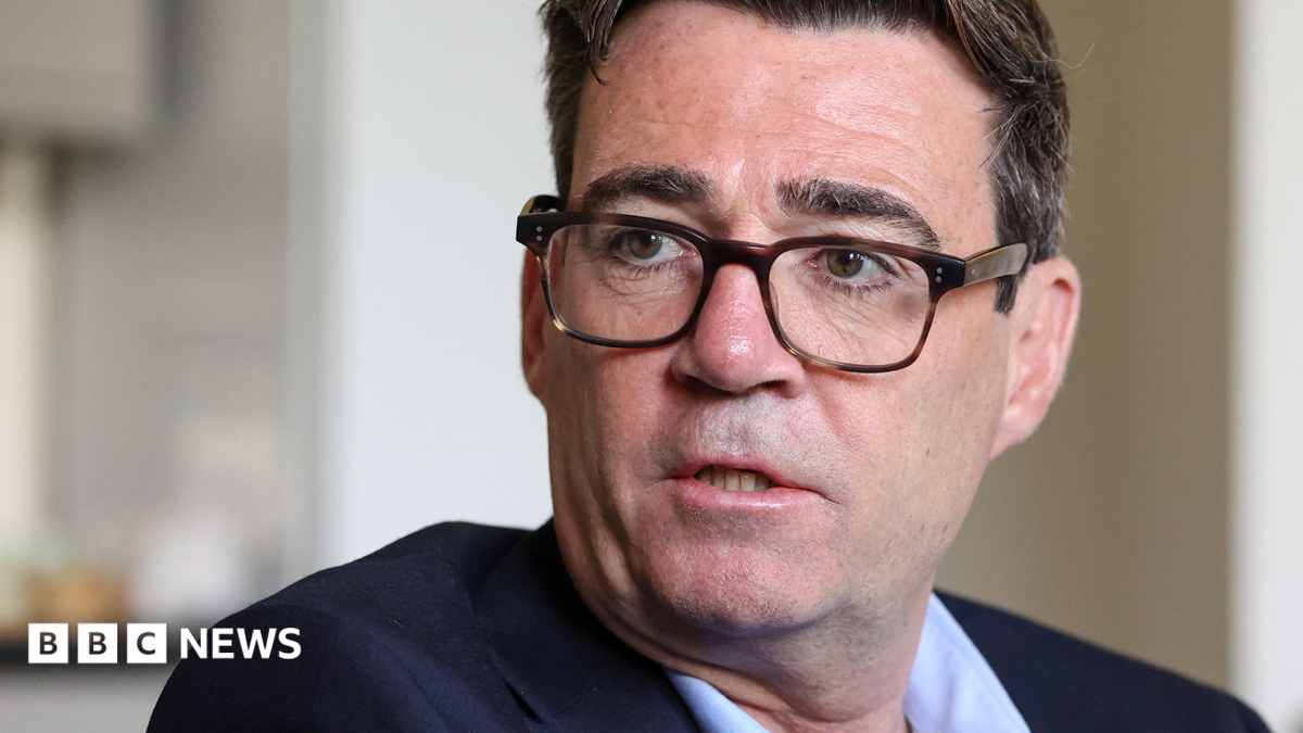 Andy Burnham is wearing a dark-coloured jacket, light blue shirt and glasses. He has short black hair.