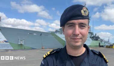 Lieutenant commander Dominic Jacob has his Royal Navy uniform on. The large grey ship with green detailing is docked in the background.