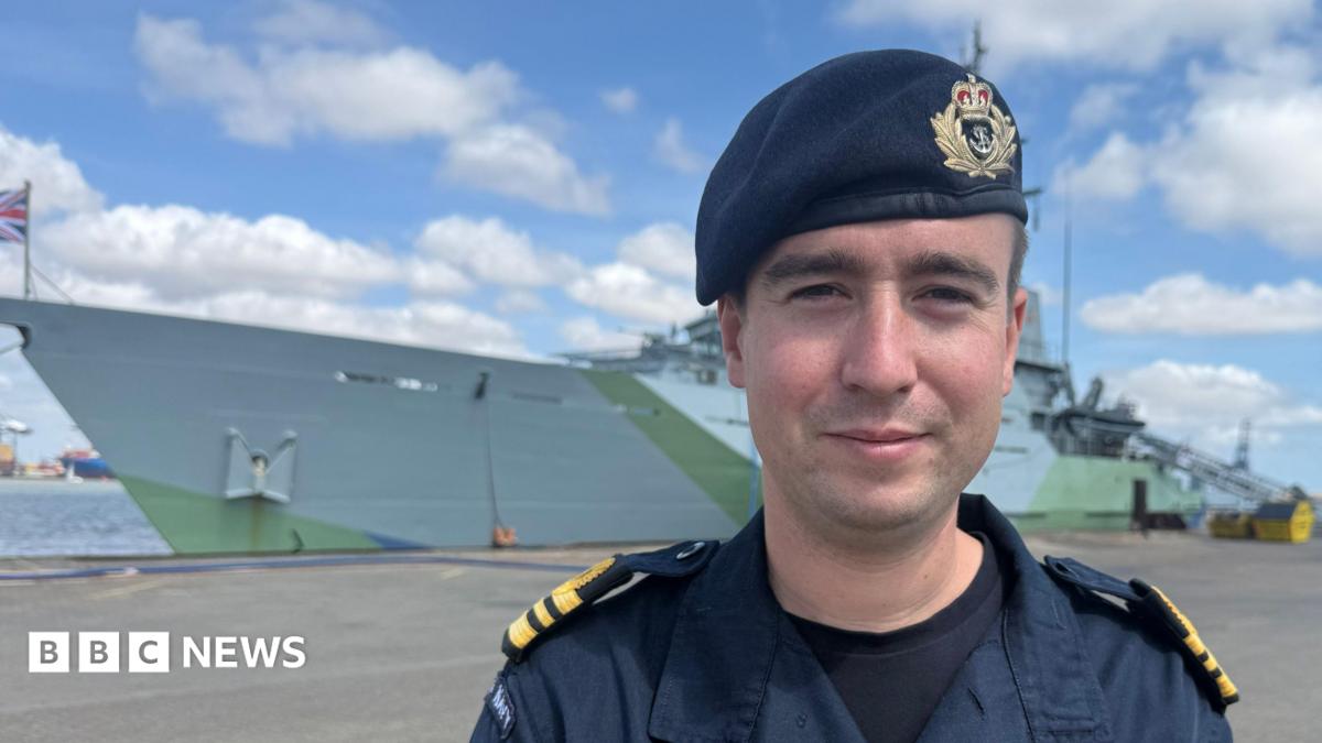 Lieutenant commander Dominic Jacob has his Royal Navy uniform on. The large grey ship with green detailing is docked in the background.