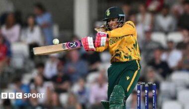 Suné Luus looks to crack the white cricket ball through the on-side, wearing green and gold South Africa kit and red and white batting gloves, with shoulder length blonde hair pretruding from bottom of her helmet