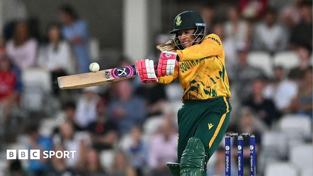 Suné Luus looks to crack the white cricket ball through the on-side, wearing green and gold South Africa kit and red and white batting gloves, with shoulder length blonde hair pretruding from bottom of her helmet