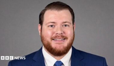 A man with short, coiffed hair, and a short ginger beard is smiling at the camera. He is wearing a blue suit and tie.