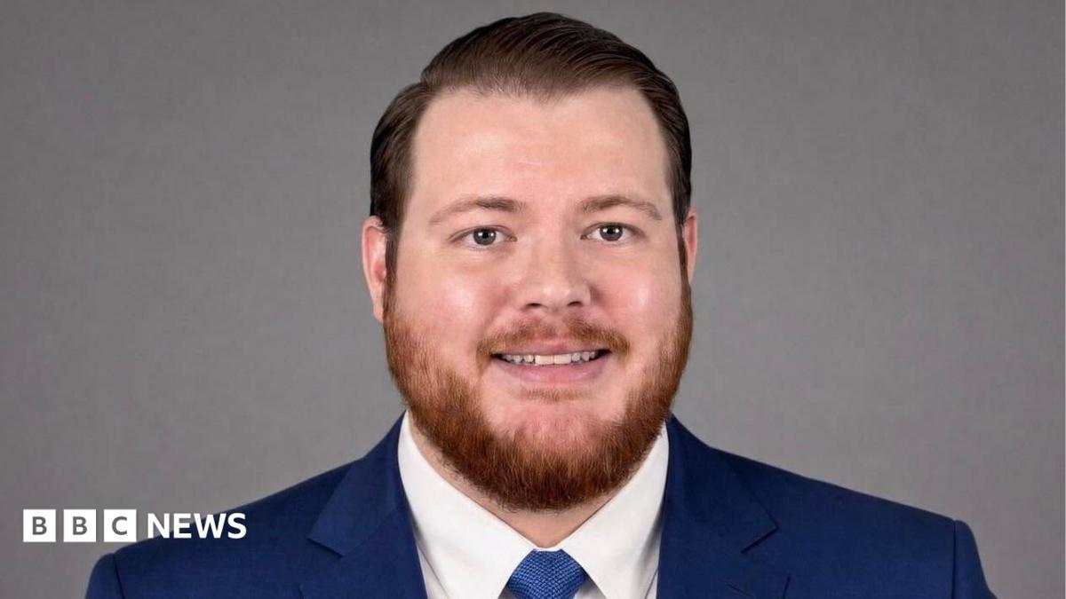 A man with short, coiffed hair, and a short ginger beard is smiling at the camera. He is wearing a blue suit and tie.