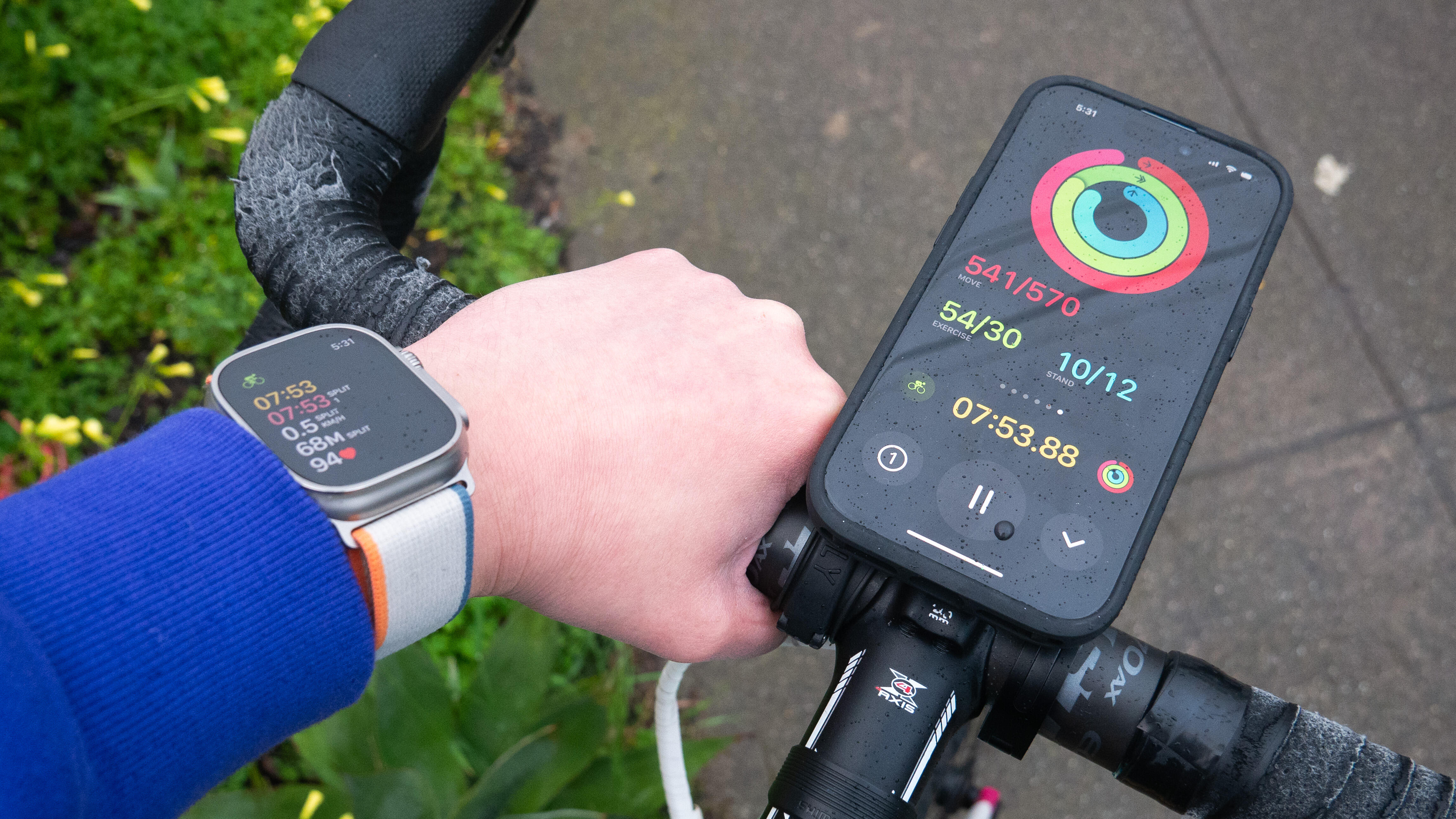 The iPhone 15 mounted on a bike, with a hand grasping the handlebar, an Apple Watch Ultra on the wrist