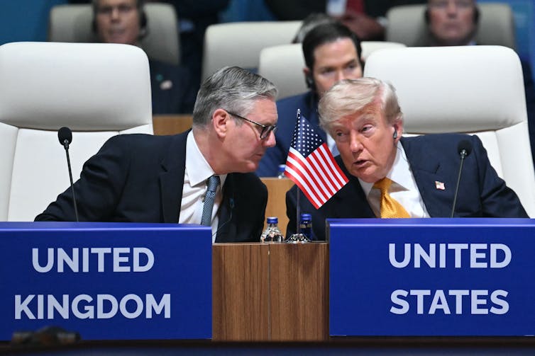 Two men in suits talk in front of signs that say United Kingdom and United States.