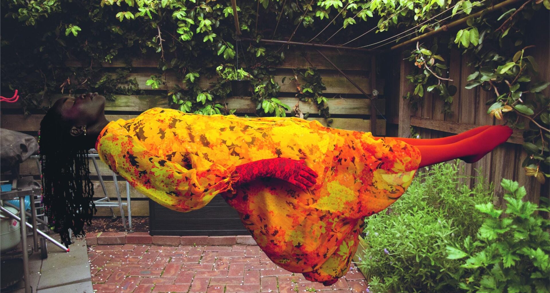 a photograph by Atong Atem of a woman wearing a warm-hued floral dress levitating as she lays horzontally