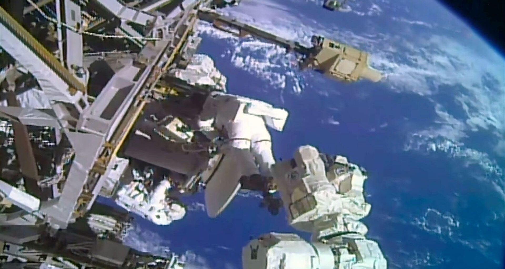 two astronauts in white spacesuits work near a large antenna dish outside a space station with a blue and white planet as their backdrop