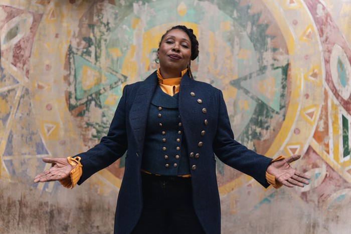 522036,TITLE:Doctor Who: Unleashed S2 Jo Martin as the Fugitive Doctor in Doctor Who stands with arms open, wearing a dark blue, double-breasted coat over a mustard high-neck blouse, posed against a weathered, circular mural painted with geometric symbols and muted earth tones.