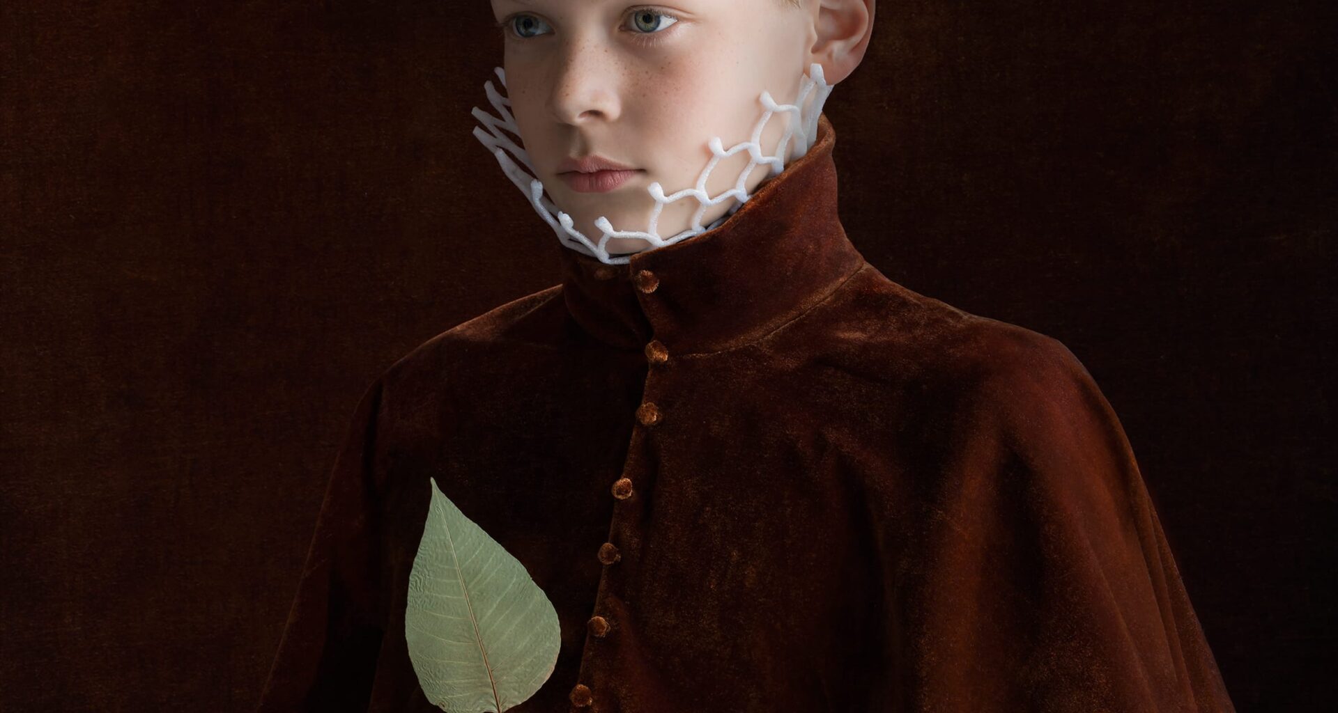 a portrait by Suzanne Jongmans of a young boy wearing a velvet jacket, with a neck ruff made of plastic used to protect fruit, holding a green leaf