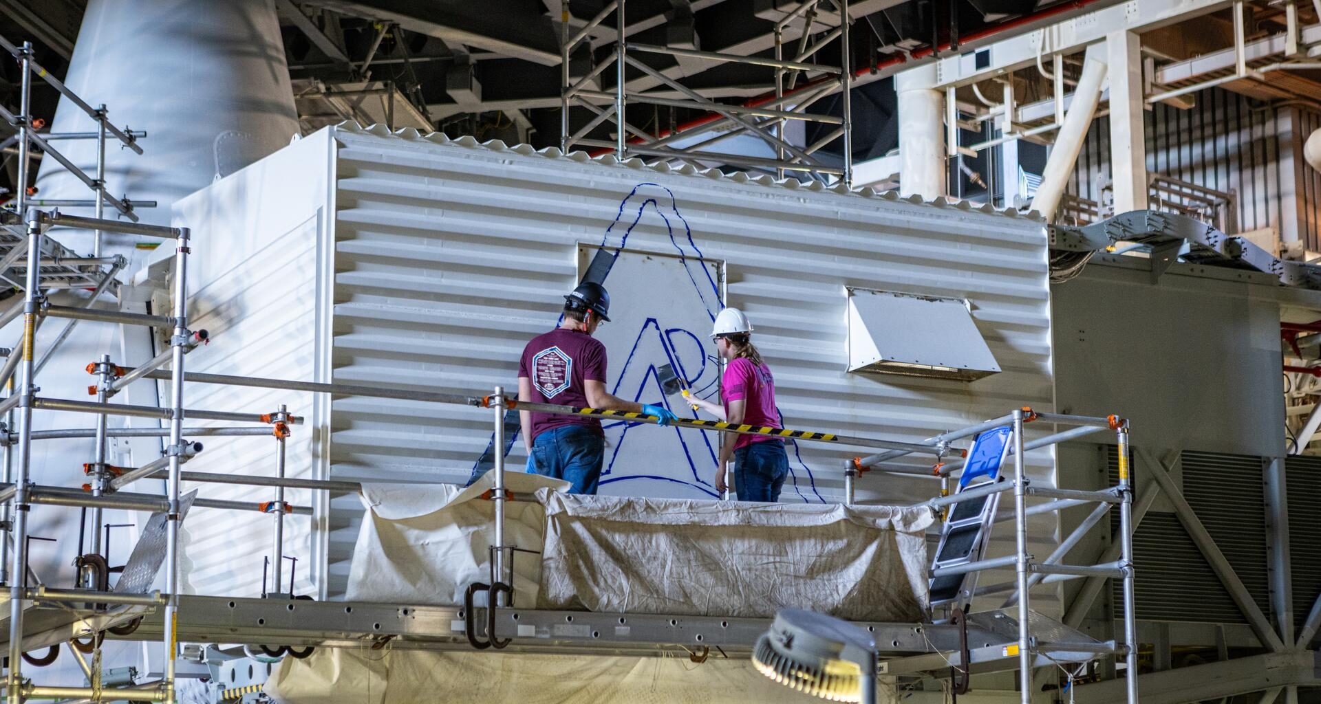 Two people standing atop scaffolding wearing hard hats. They are facing a large white rectangular structure that has an outline of the Artemis logo in blue. The two are preparing to paint the logoSean McCrary and Katie Mortensen, mechanical engineering technicians, paint NASA’s Artemis logo on the White Room connected to the crew access arm and mobile launcher inside the Vehicle Assembly Building at NASA’s Kennedy Space Center in Florida on Thursday, Dec. 4, 2025.