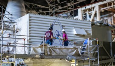 Two people standing atop scaffolding wearing hard hats. They are facing a large white rectangular structure that has an outline of the Artemis logo in blue. The two are preparing to paint the logoSean McCrary and Katie Mortensen, mechanical engineering technicians, paint NASA’s Artemis logo on the White Room connected to the crew access arm and mobile launcher inside the Vehicle Assembly Building at NASA’s Kennedy Space Center in Florida on Thursday, Dec. 4, 2025.