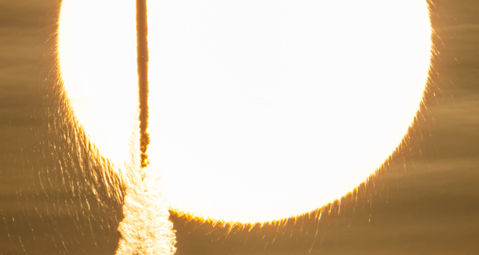 In a sepia-toned photo, the Sun appears as a bright, enormous orb that appears to have frayed edges. Against this backdrop, a tall and slender rocket is seen in silhouette atop a cloudy plume.