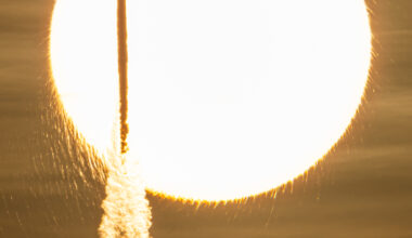 In a sepia-toned photo, the Sun appears as a bright, enormous orb that appears to have frayed edges. Against this backdrop, a tall and slender rocket is seen in silhouette atop a cloudy plume.