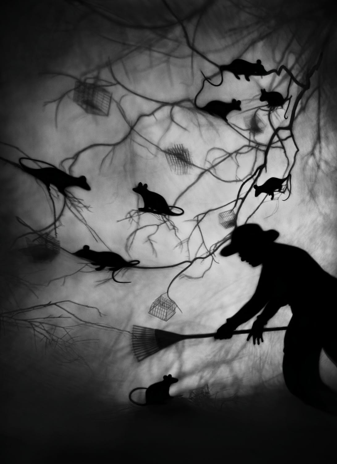 a black-and-white photograph by Susan kae Grant of a silhouetted dream-like scene of a man with a hat and a broom swiping at branches full of large rats