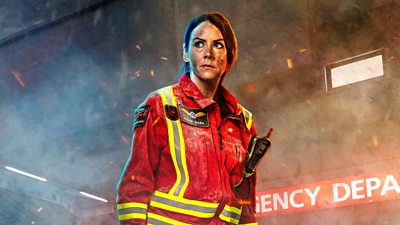 A woman in a red emergency uniform, stood by a hospital with smoke and embers around her