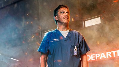 A man in blue medical scrubs stood by a hospital with smoke and embers blowing around him