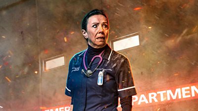 A woman in dark nay and white scrubs and stethoscope around he neck stood outside a hospital with smoke and embers around her