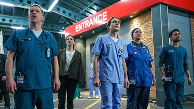 A group pf medical staff stand outside the doors of an Emergency Department looking anxiously at something happening off screen