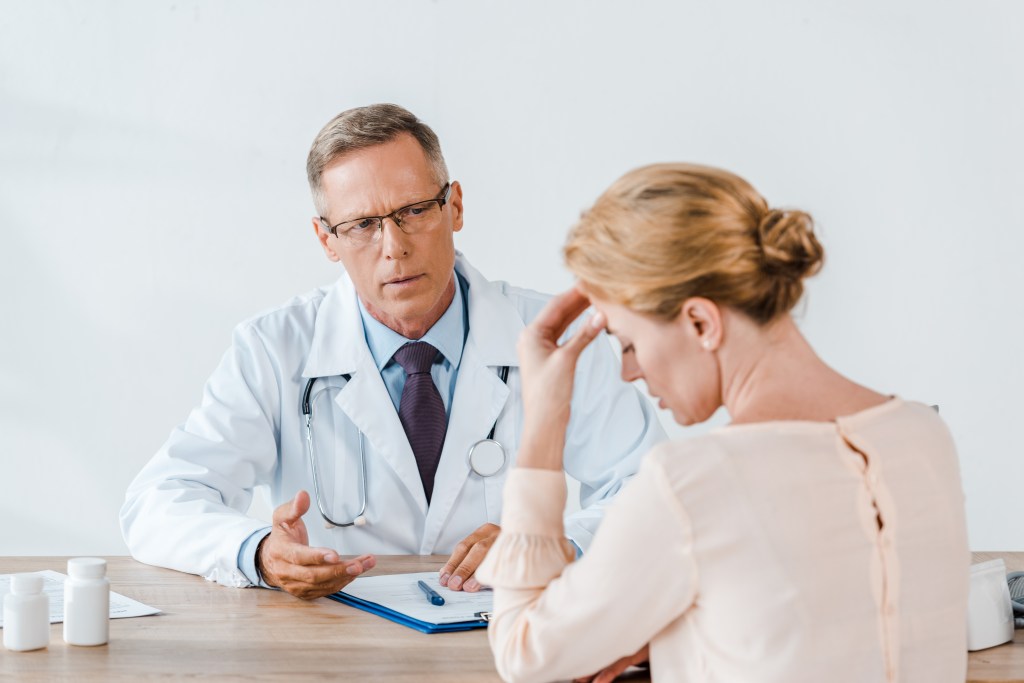 A male doctor in glasses talks to an upset female patient touching her head at a table.