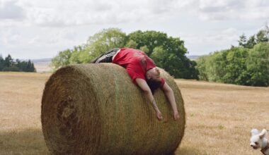 a photographic portrait of a young man in a field, laying on his back on top of a large hay bale, with a small white dog energetically running into the frame at the bottom right