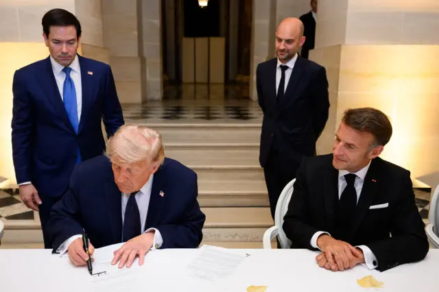 US President Donald Trump (sitting, left) signs his name at the bottom of the Memorandum of Understanding as French President Emmanuel Macron (sitting, right) observes. They're both wearing black suits. Behind them, standing, are US Secretary of State Marco Rubio and French Foreign Minister Jean-Noel Barrot