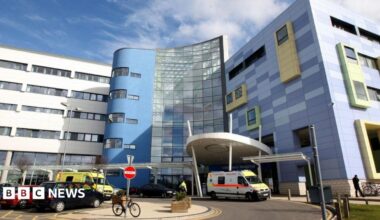 A picture of the John Radcliffe Hospital in Oxford. It is blue and grey, are there are cars and ambulances parked in front of it.