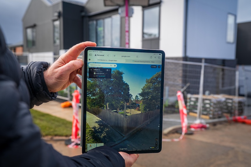 A man, standing in front of a townhouse construction site, holds up a photo on an iPad screen