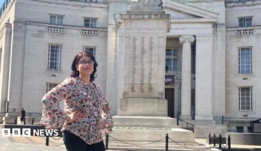 Tahmina Saleem stands in a paved square in front of a large stone building in Luton and a war memorial, with blue sky and clouds overhead. She is wearing black trousers and a patterned blouse.