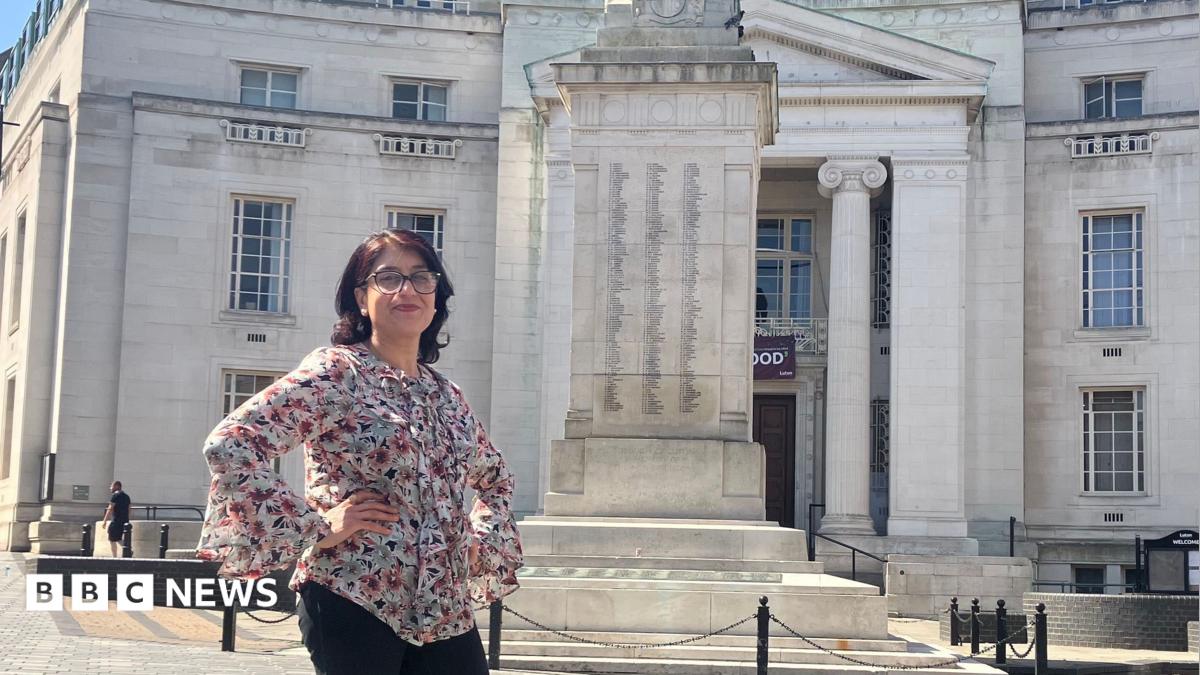 Tahmina Saleem stands in a paved square in front of a large stone building in Luton and a war memorial, with blue sky and clouds overhead. She is wearing black trousers and a patterned blouse.