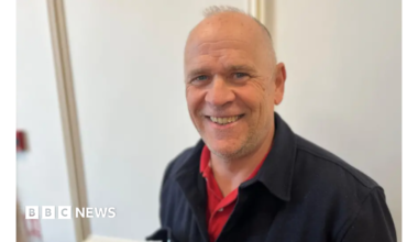 Mark Laine has short grey hair and is wearing a black jacket over a red shirt