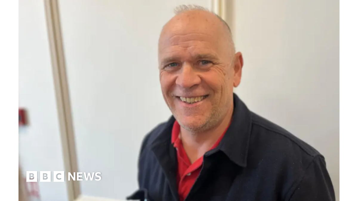 Mark Laine has short grey hair and is wearing a black jacket over a red shirt