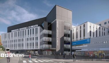 Modern hospital building exterior with multiple storeys, a central entrance tower, and people crossing the street in front. Visible signage reads “University Hospitals Plymouth” and “NHS”, under a clear blue sky.