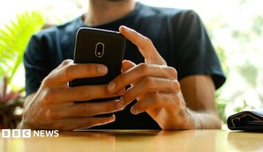A man, whose face has been cut off in the picture, holding a phone with both hands while resting his arms on a wooden table. There are out-of-focus plants in the background. He's wearing a dark green t shirt.