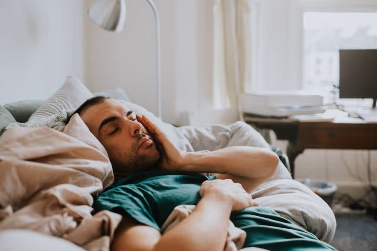 A man has just woken up in a domestic environment, either a living room or a bedroom. He is cozy in duvets and pillows. He rubs his eyes and face sleepily as he comes to.
