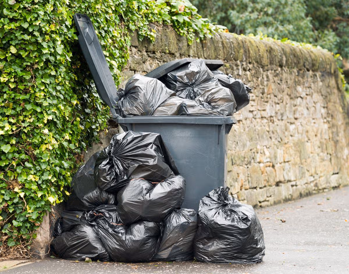 A full wheelie bin with rubbish bags overflowing onto the pavement