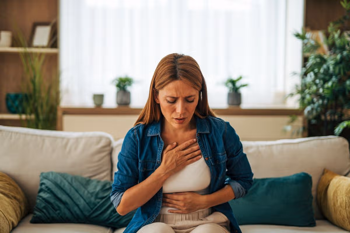Mid adult woman experiencing sudden chest pain, shortness of breath and discomfort while sitting on the sofa in her living room, possible symptoms of heart attack, angina or other medical emergency