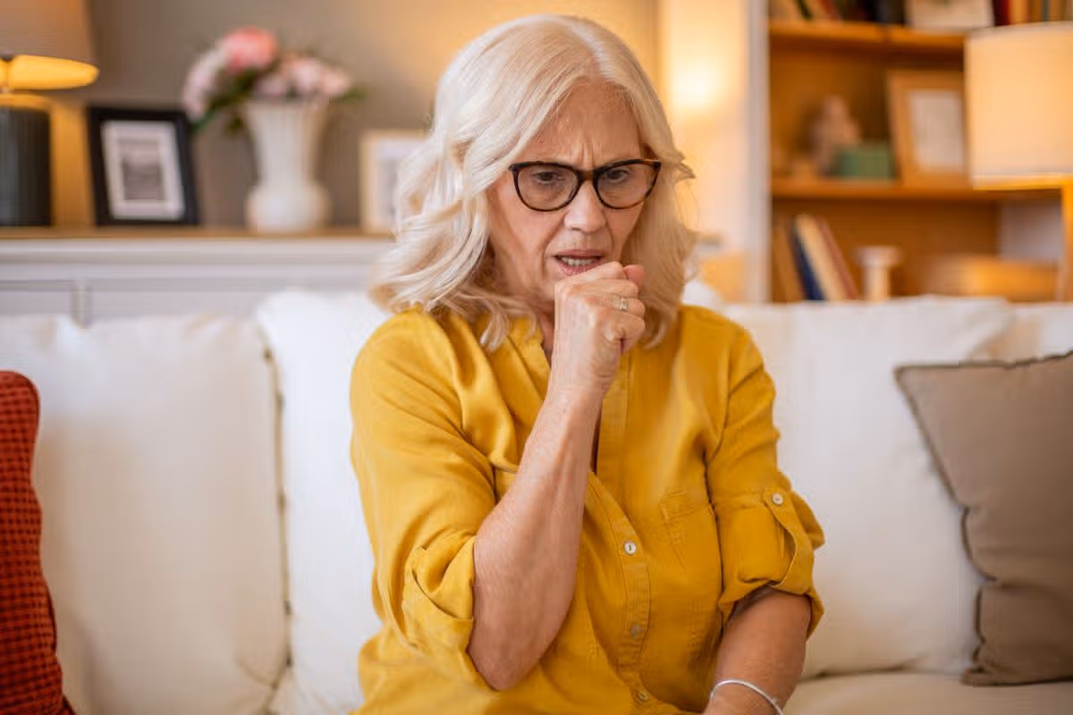 Senior woman coughing while sitting on a sofa at home, suffering from a cold or flu